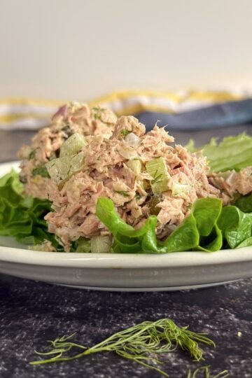 tuna salad with celery, shallots, and fresh dill stacked on leaf lettuce on a white plate. a few pieces of fresh dill in the front.