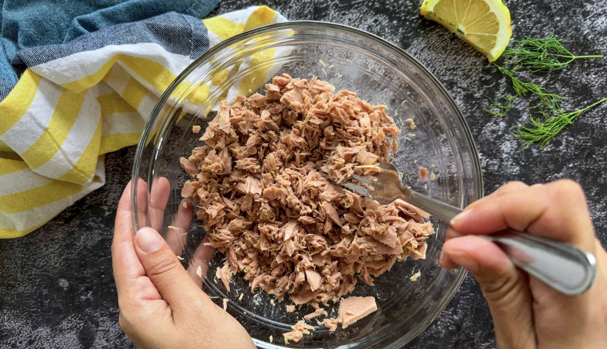 hand with a fork flaking canned tuna in a bowl. lemon wedge and fresh dill in the background.