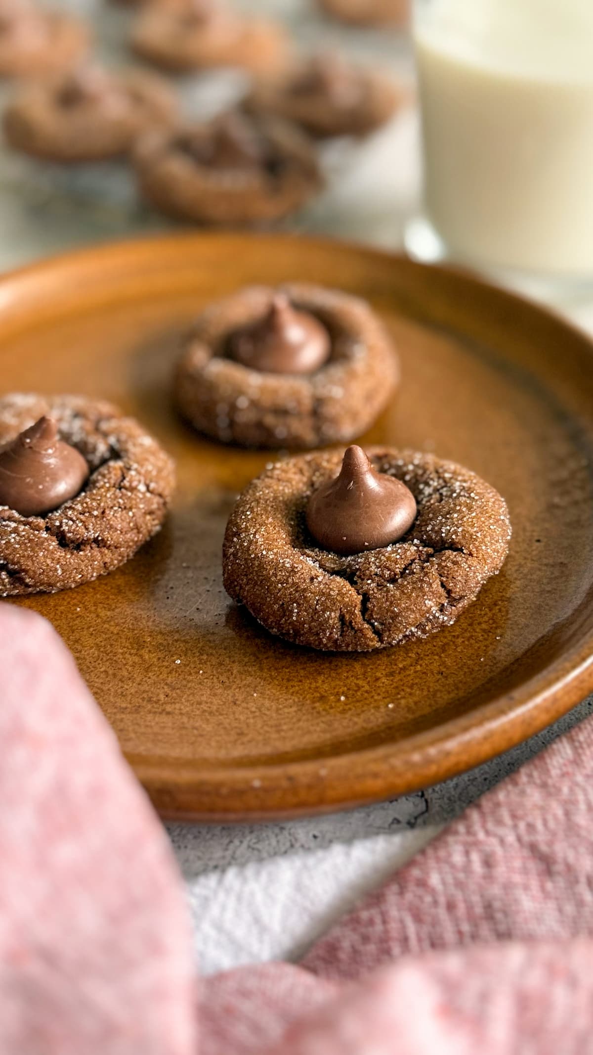 3 hershey kiss gingerbread cookies sitting on a brown plate. More in the background next to a glass of milk.