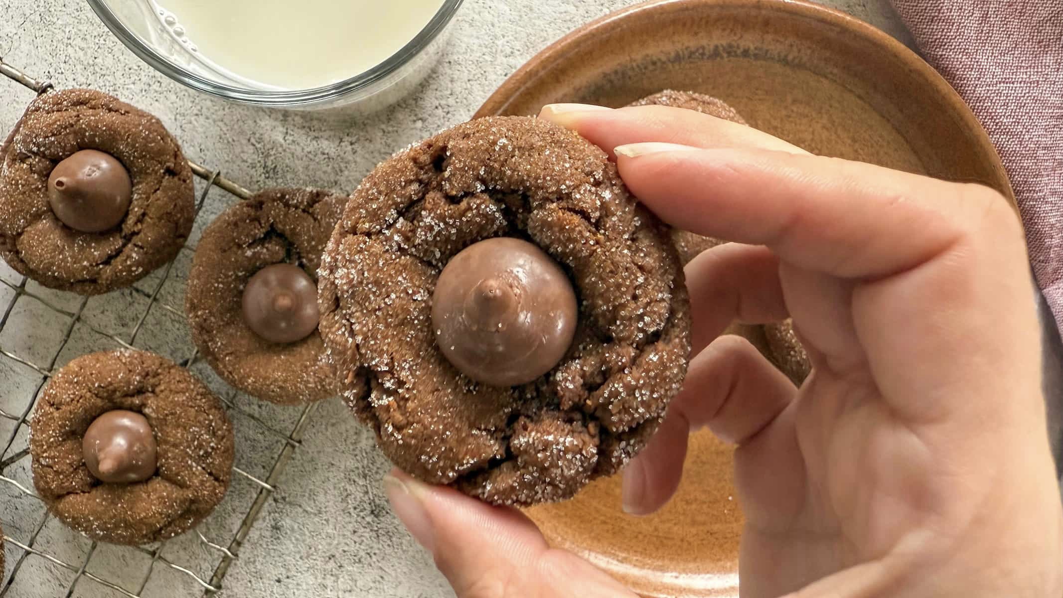hand holding a gingerbread kiss cookie over a wire rack with more. glass of milk next to it.