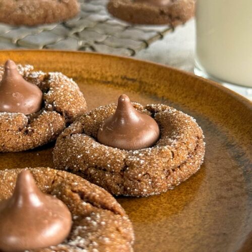 side view of brown hershey kiss cookies on a plate. More on a wire rack in the background next to a glass of milk.