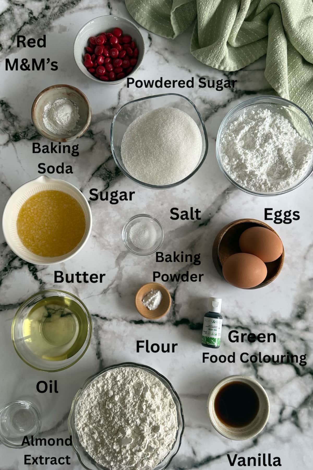 ingredients for grinch crinkle cookies laid out in separate bowls - red M&M's, powdered sugar, white sugar, butter, oil, eggs, baking soda, baking powder, salt, green food colouring, almond extract, vanilla, and flour.
