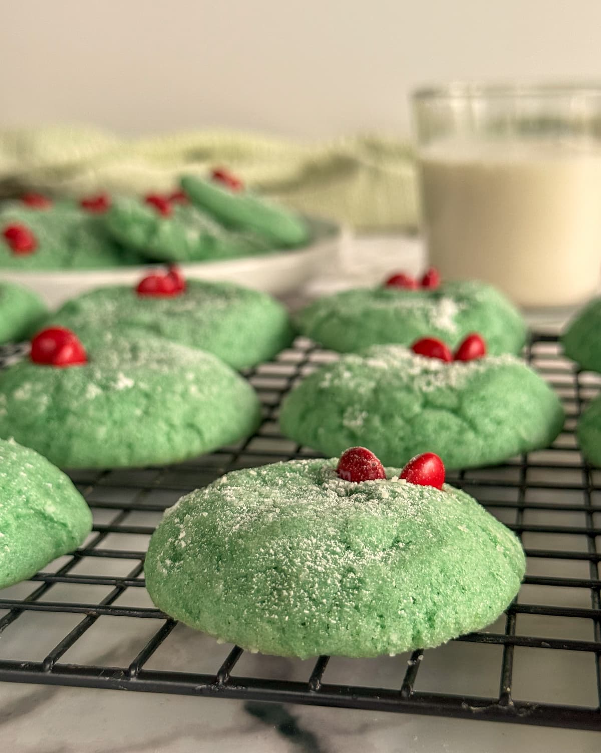 grinch crinkle cookies with a red heart and powdered sugar on a wire rack. more cookies in a bowl next to a glass of milk in the background.