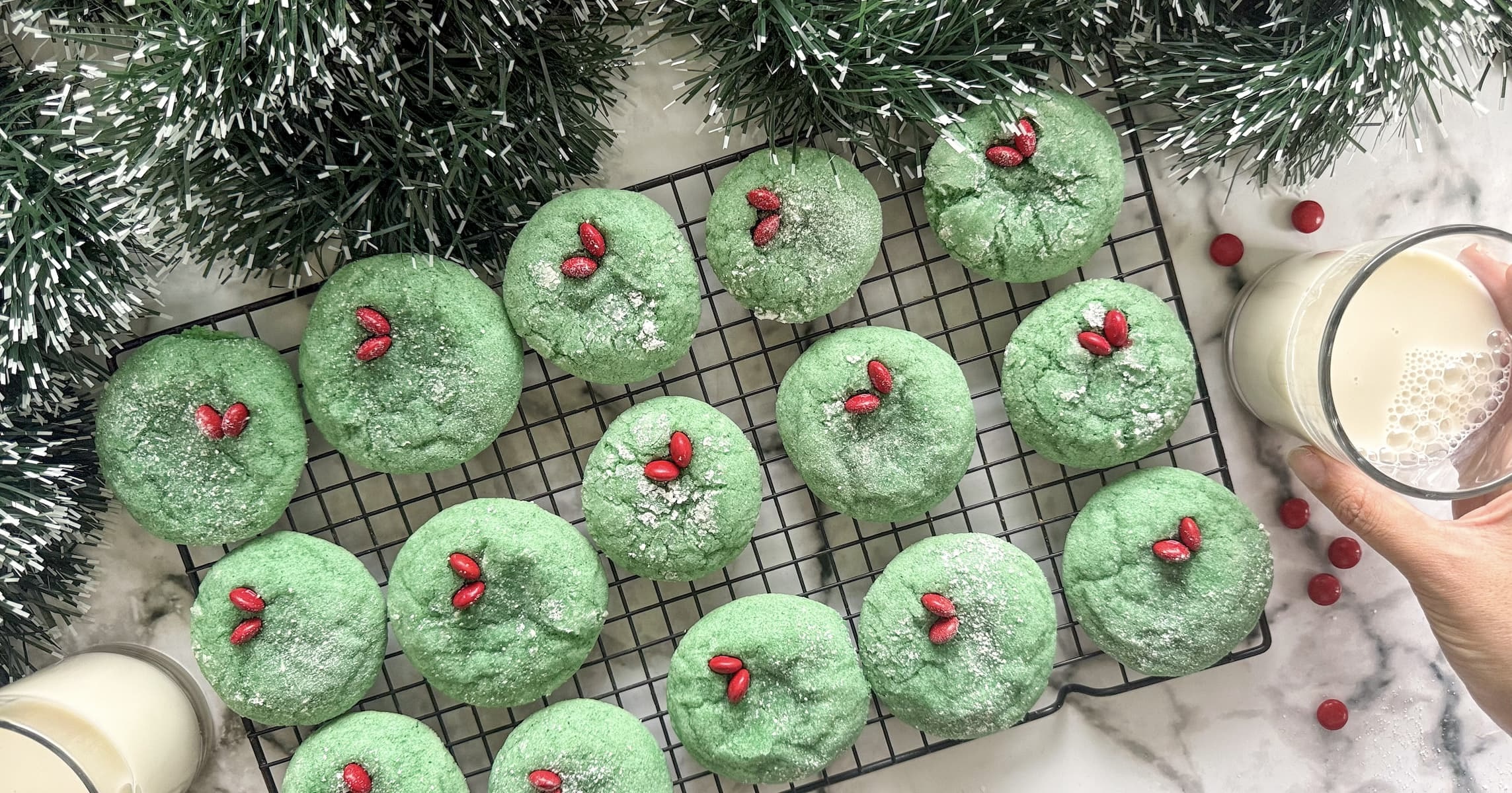 grinch crinkle cookies laid out on a wire rack next to two glasses of milk. hand reaching for one of the glasses.