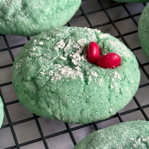 close up of a green crinkle cookie with a red M&M heart on a wire rack surrounded by more cookies.