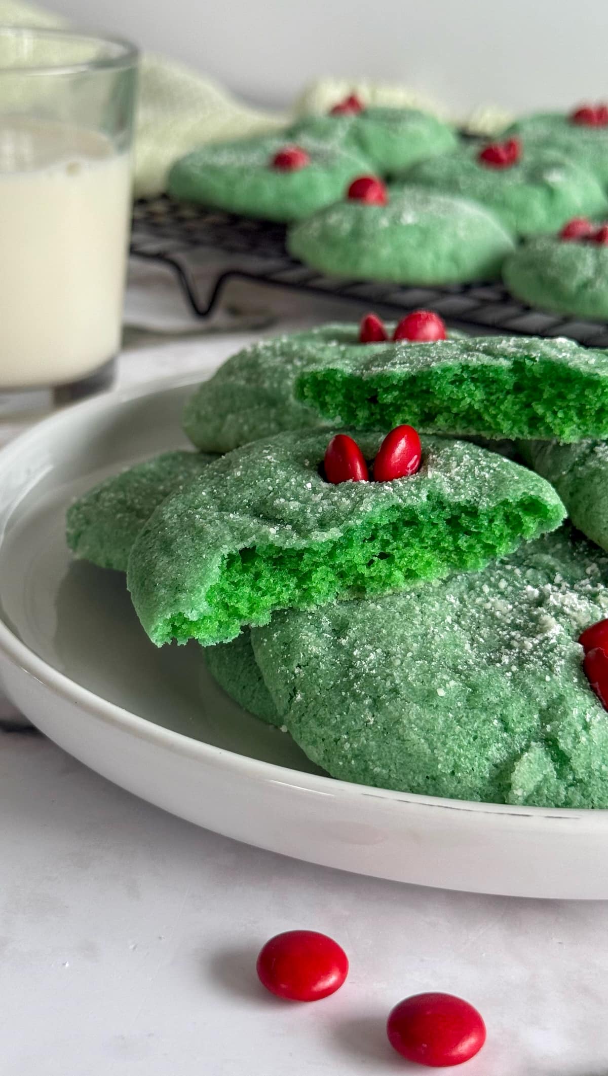 halved green grinch cookie on a plate with more. more cookies on a wire rack in the background next to a glass of milk.