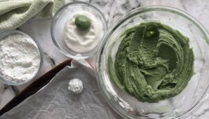bowl of powdered sugar next to a bowl of granulated sugar with a green ball of dough on top. bowl of green batter and parchment lined baking sheet with a sugar coated dough ball on the baking sheet.