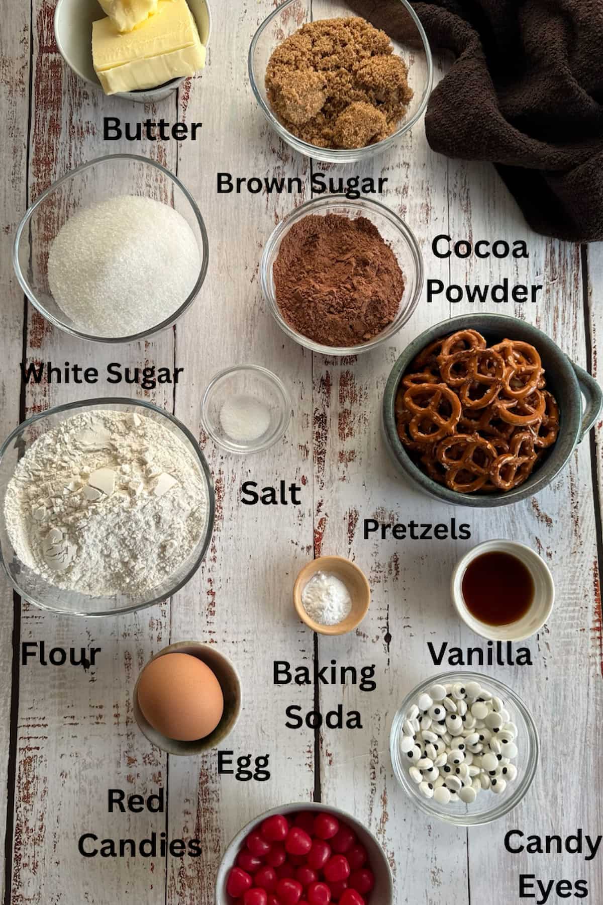 ingredients for reindeer christmas cookies set out in separate bowls - butter, brown and white sugar, cocoa powder, red candies, pretzels, candy eyes, salt, egg, baking soda, vanilla, and flour.