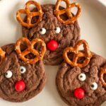 Reindeer Christmas cookies with pretzel antlers and red noses on a white plate.