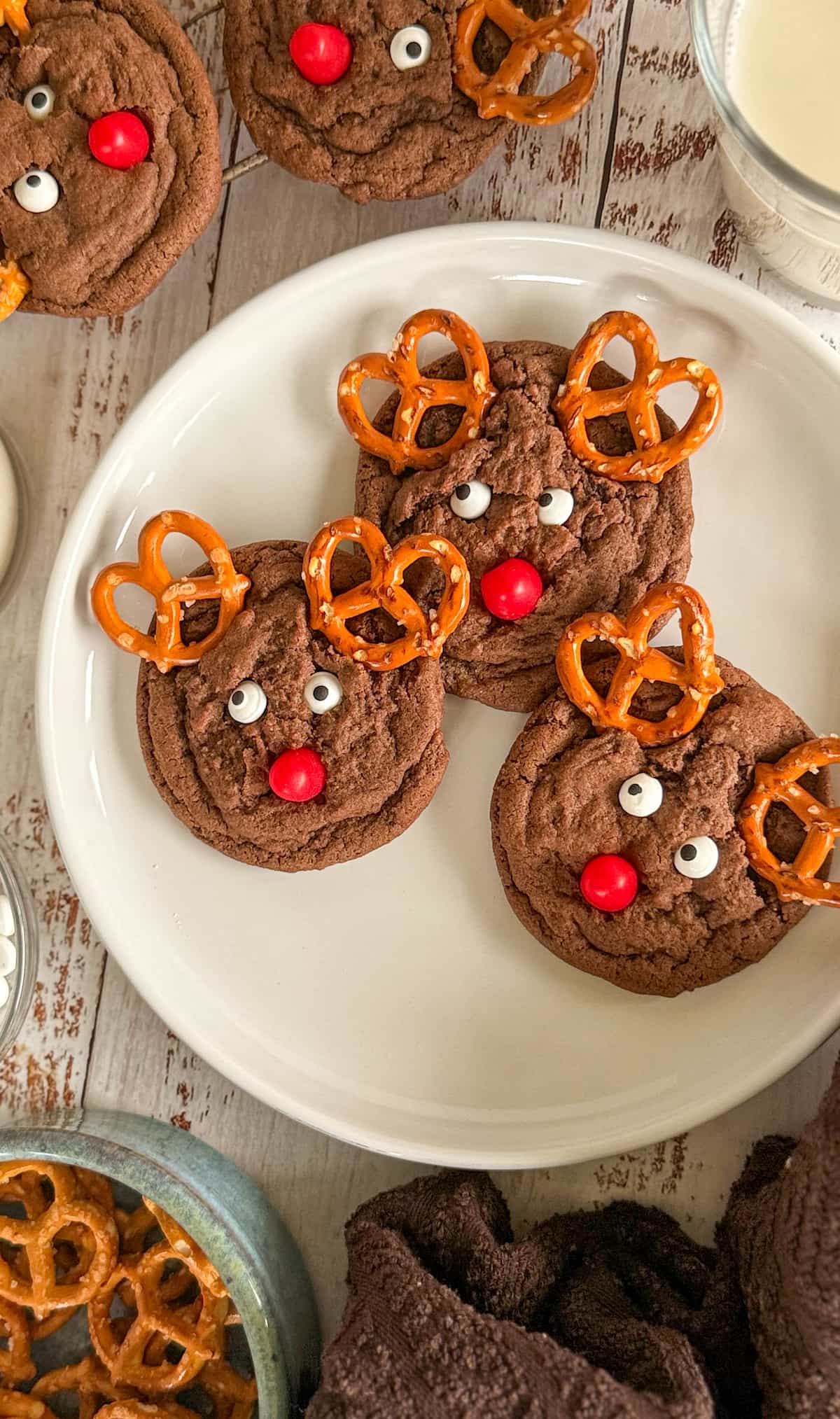 white plate with 3 reindeer christmas cookies. more cookies, a glass of milk, and a bowl of pretzels in the background.