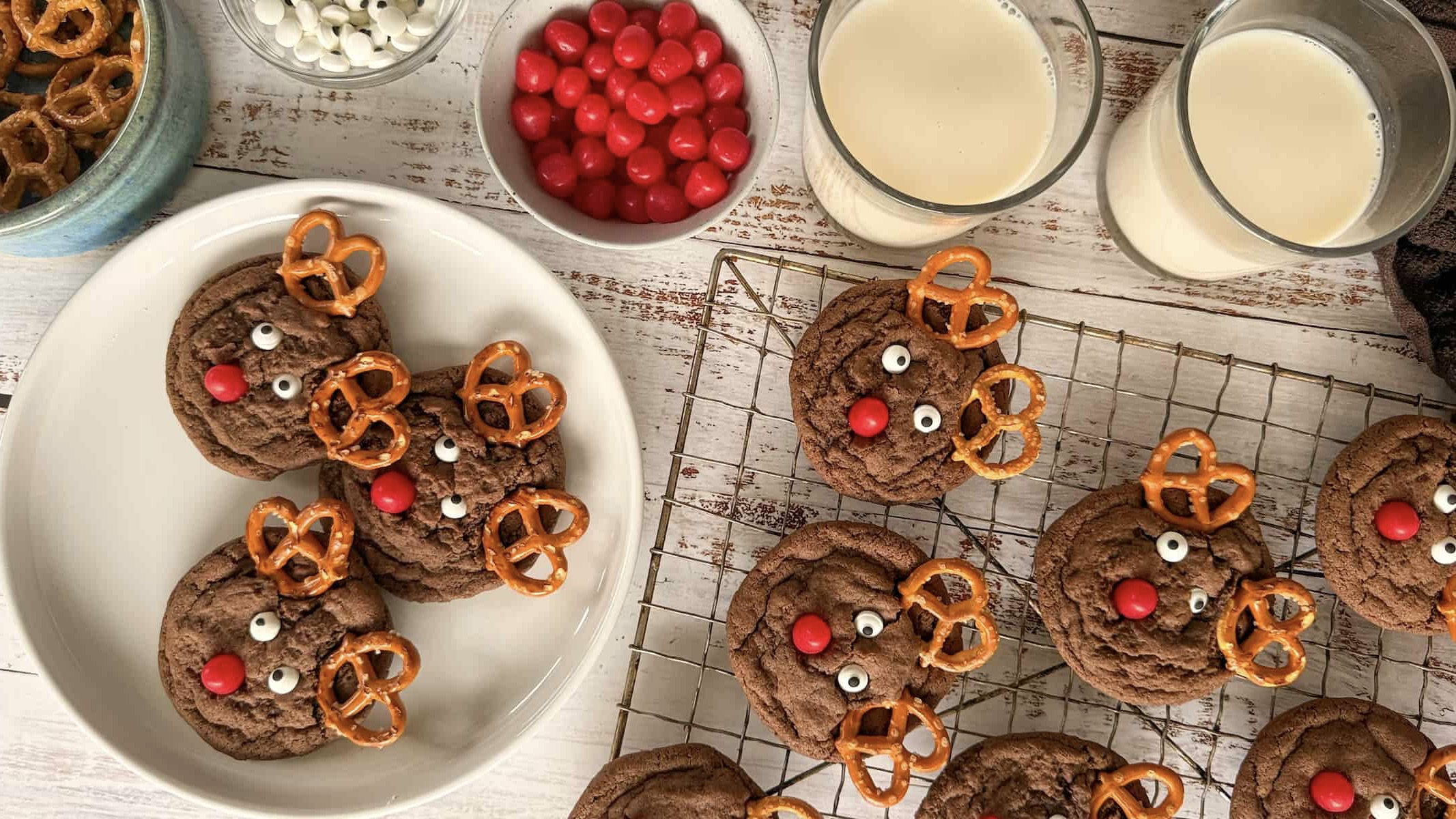several reindeer christmas cookies on a wire rack next to a white plate with 3 more cookies. 2 glasses of milk, a bowl of red candy noses, candy eyes, and pretzels next to it.