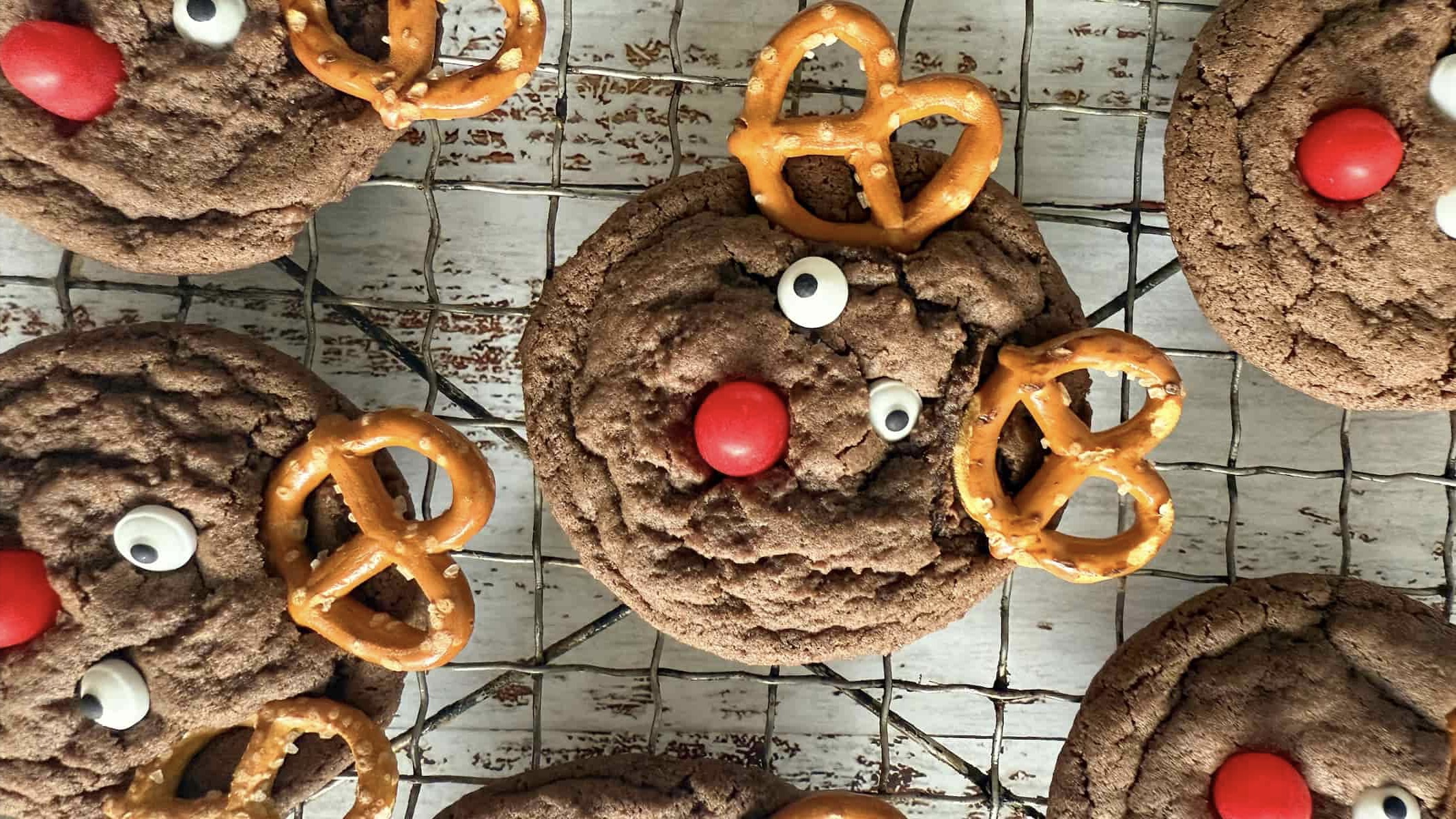 close up of reindeer christmas cookies with pretzel antlers, candy eyes, and a red candy nose on a wire rack.