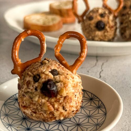 mini cheese ball with pretzel antlers, a dried cranberry nose, and black peppercorn eyes. more on a plate in the background next to some crostini.