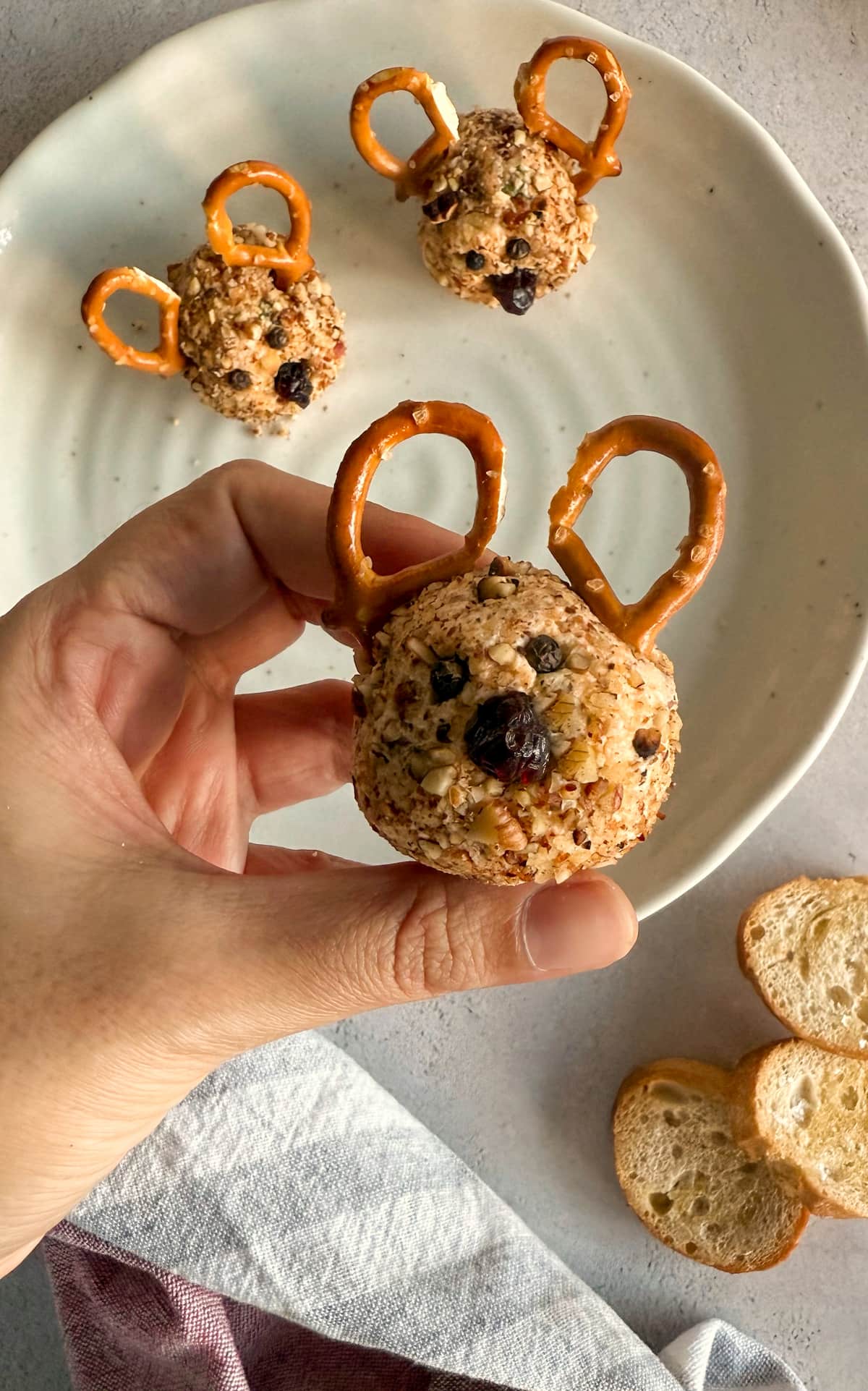 hand holding a mini reindeer cheese ball with pretzel antlers, black peppercorn eyes, and a dried cranberry nose, over a plate with more.
