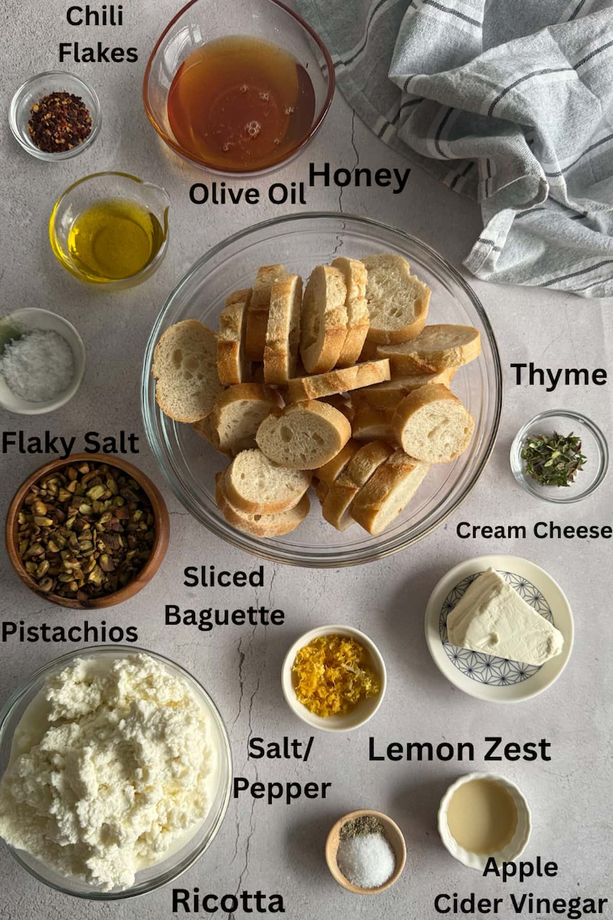 ingredients for ricotta and hot honey crostini set out in separate bowls - chili flakes, honey, olive oil, flaky salt, thyme, sliced baguette, cream cheese, ricotta, salt, pepper, apple cider vinegar, lemon zest, and pistachios.