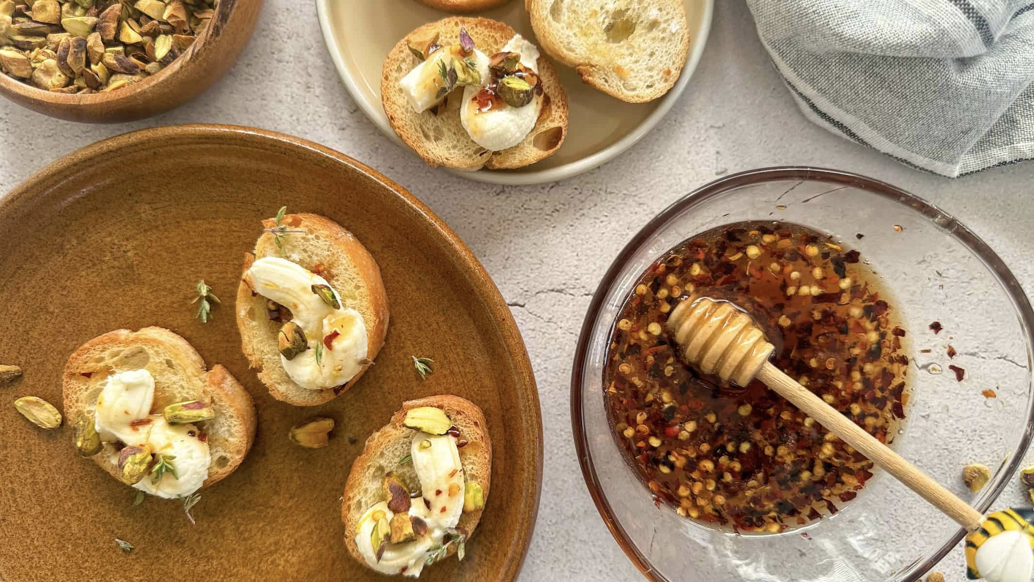 bowl of hot honey with a honey dipper next to 2 different plates of ricotta and hot honey crostini. small bowl of chopped pistachios next to the plate.