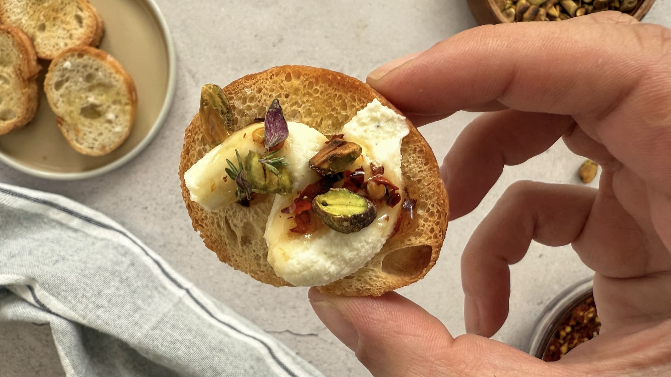 hand holding a sliced baguette with whipped ricotta, chopped pistachios, and hot honey. plate with 3 plain crostini in the background.