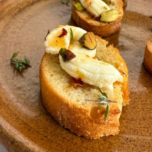 2 pieces of sliced toasted baguette (crostino) topped with a swirl of whipped ricotta, chopped pistachios, hot honey, and thyme on a brown plate.