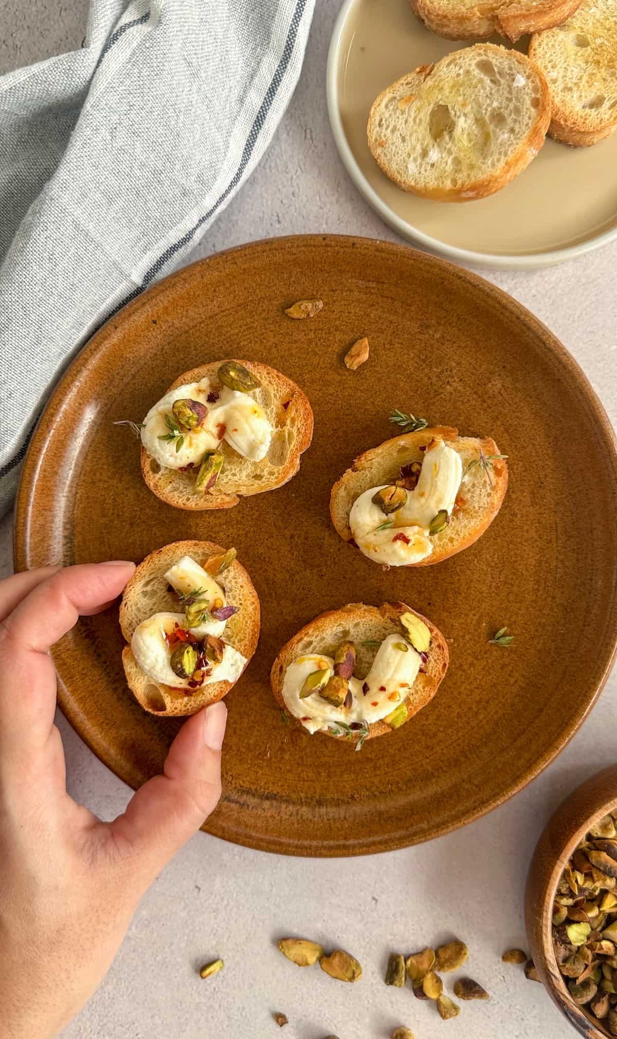 hand grabbing a crostini topped with chopped pistachios, thyme, ricotta and hot honey, off a plate with 4 altogether. another plate in the background of plain crostini as well as a bowl of chopped pistachios.
