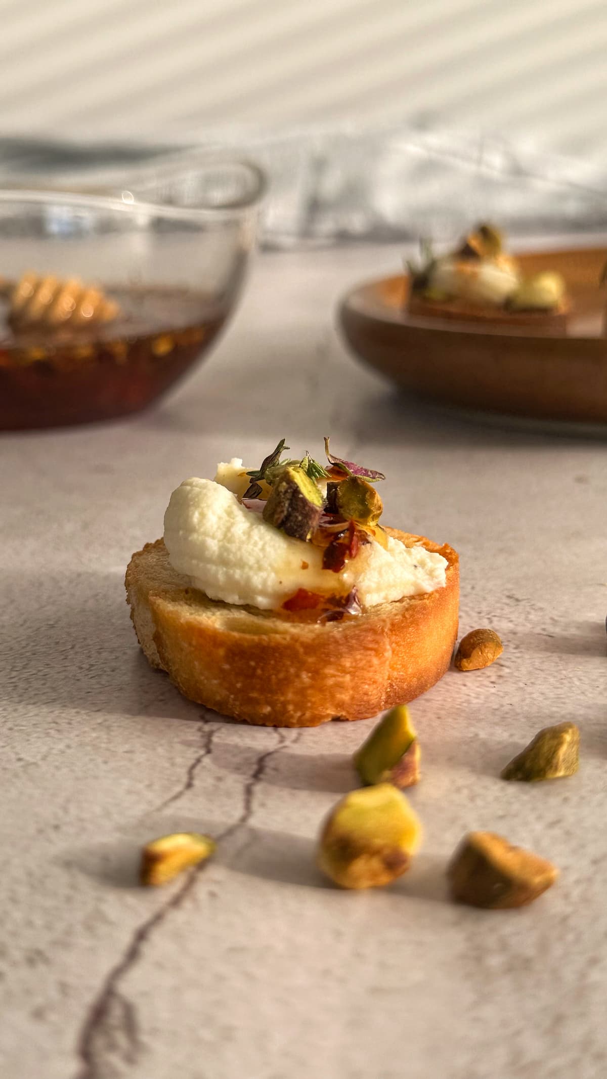 single crostino topped with ricotta, hot honey, pistachios, and thyme next to some scattered pistachios. bowl of hot honey and plate of more crostini in the background.