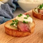 steak crostini with horseradish cream and chives on a board next to another crostini and a bowl of more horseradish cream.