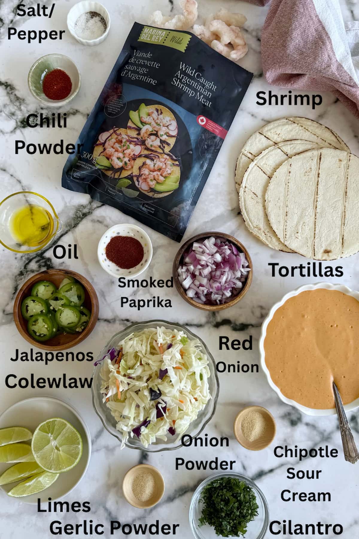 ingredients for air fryer shrimp tacos laid out - salt/pepper, chili powder, smoked paprika, garlic powder, onion powder, oil, shrimp, tortillas, coleslaw, red onion, cilantro, limes, jalapenos, chipotle sour cream.