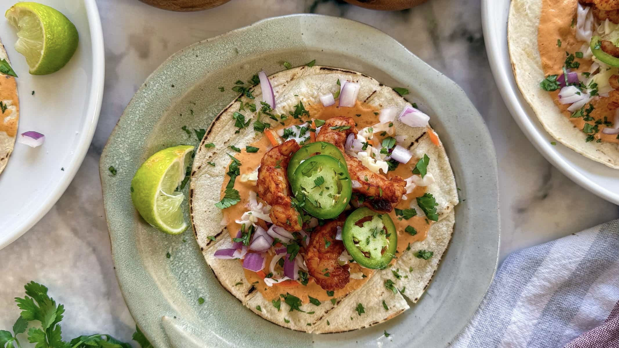 loaded shrimp taco on a plate with chopped red onions, cilantro, sliced jalapeno, lime, and a bright orange sauce. Surrounded by two more plates of tacos.