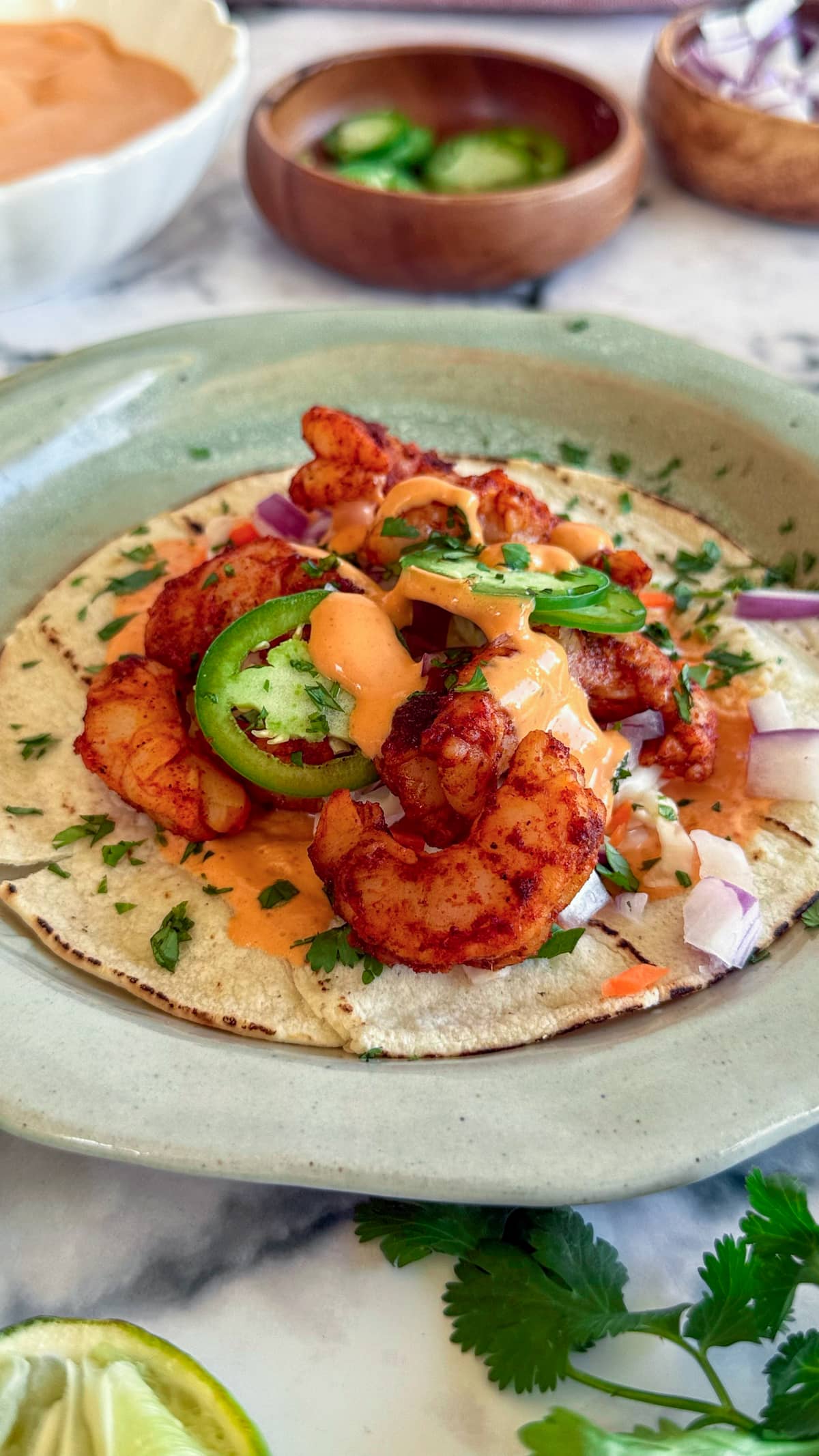 side view of a seasoned shrimp taco on a plate topped with sliced jalapeno, red onion, cilantro, and a bright orange sauce.