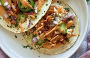 two air fryer shrimp tacos on a plate with jalapeno, red onion, cilantro, and a creamy bright orange sauce.