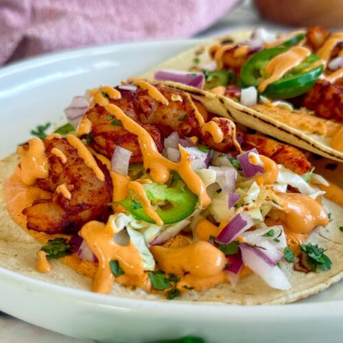 side view of two shrimp tacos on a plate with red onions, jalapenos, cilantro, and a bright orange chipotle sauce.