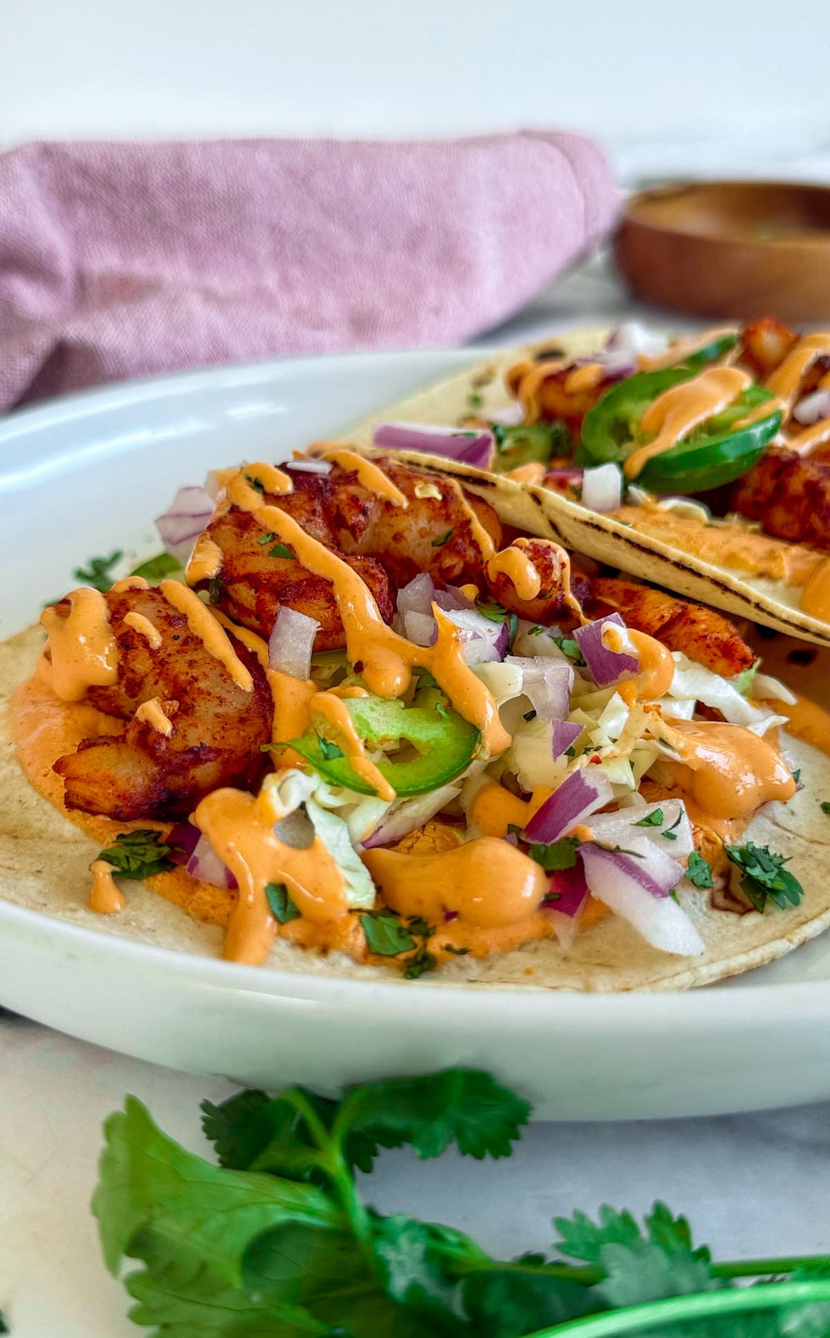 side view of two shrimp tacos on a plate with red onions, jalapenos, cilantro, and a bright orange chipotle sauce.