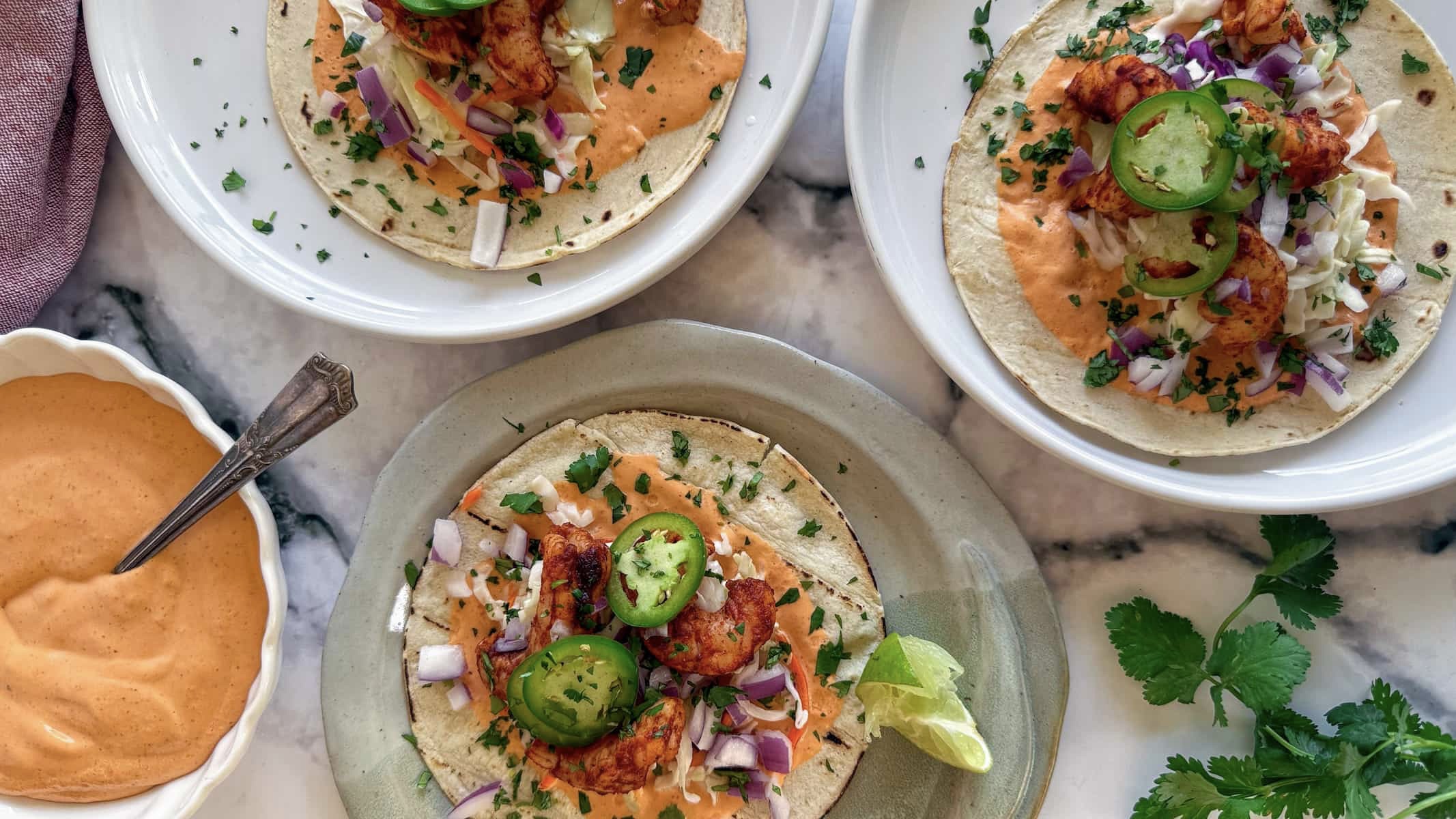 3 air fryer shrimp tacos on plates next to a creamy bright orange sauce in a bowl. Tacos are loaded with air fried shrimp, red onions, coleslaw, jalapenos, cilantro, and lime.