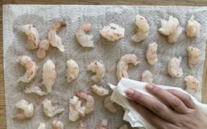 hand patting down raw pieces of shrimp with paper towel on a cutting board.