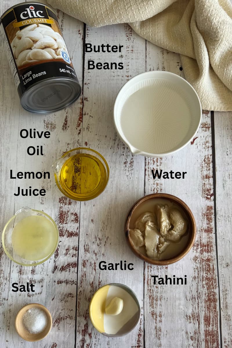 ingredients for butter bean hummus laid out in small bowls - olive oil, lemon juice, garlic, salt, tahini, water, and a can of butter beans.