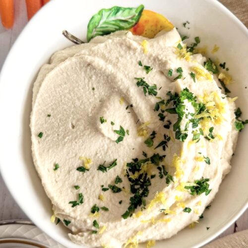 smooth butter bean hummus in a white bowl with lemon zest and chopped parsley surrounded by sliced cucumbers and baby carrots.