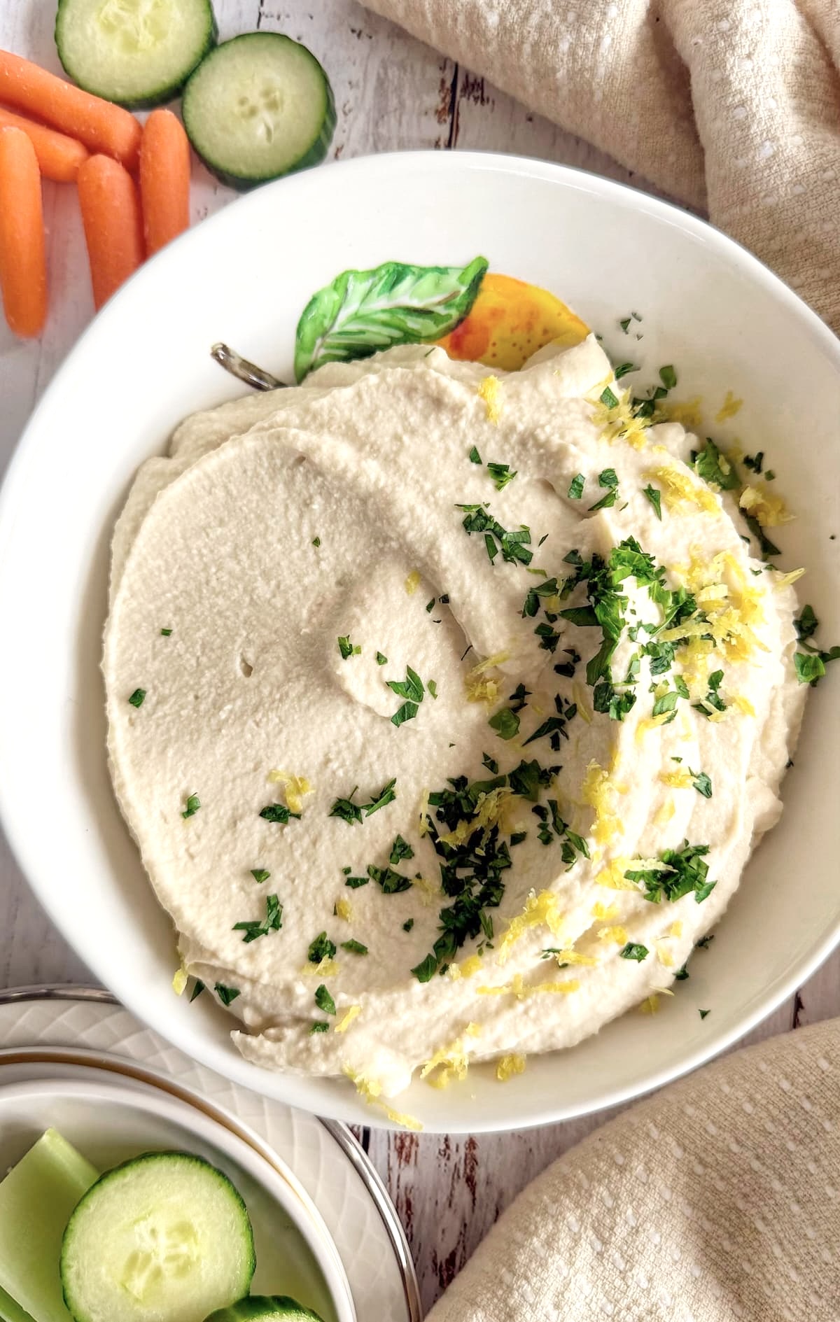 smooth butter bean hummus in a white bowl with lemon zest and chopped parsley surrounded by sliced cucumbers and baby carrots.