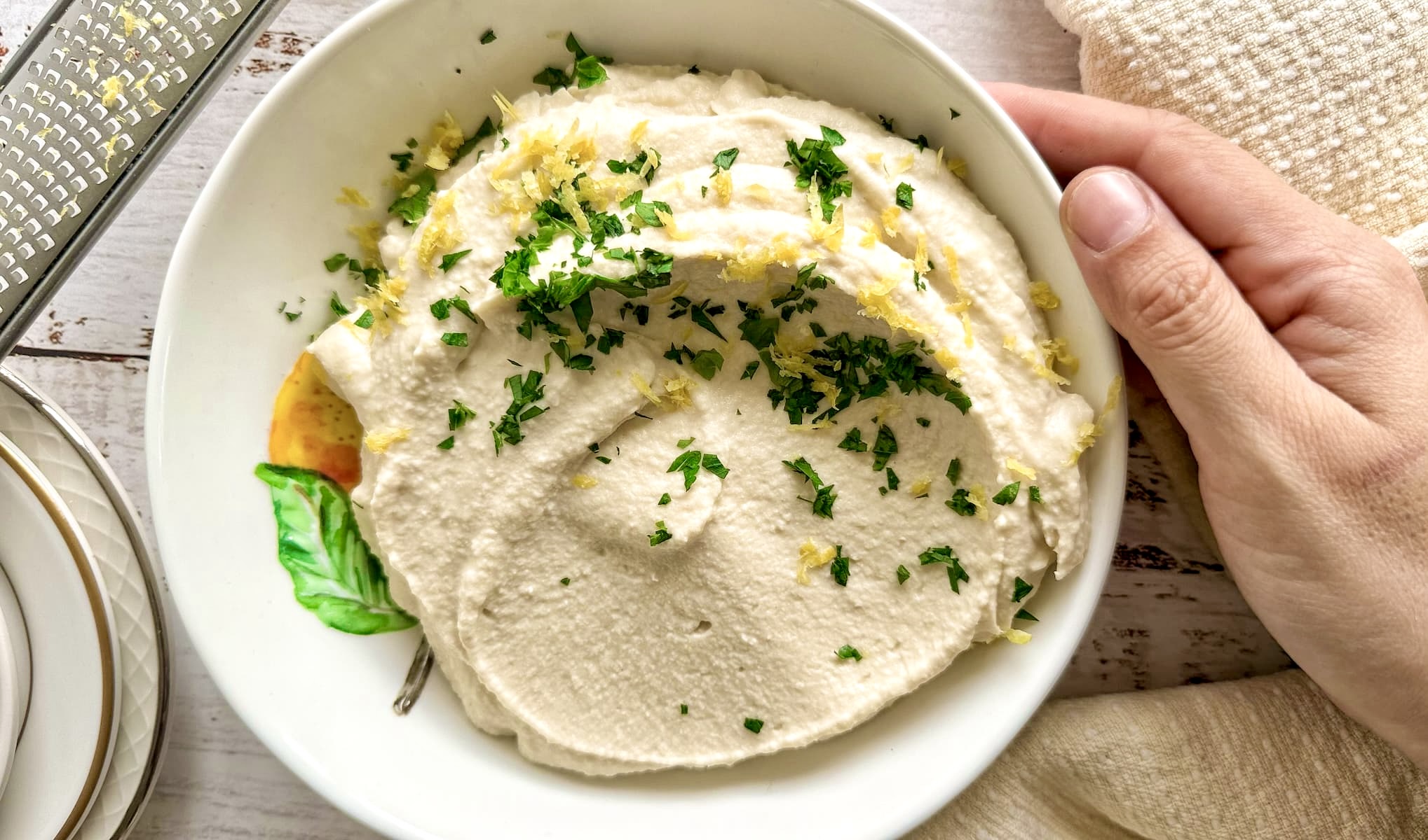 hand touching a bowl of white bean hummus with lemon zest and parsley next to a few plates and a microplane grater.