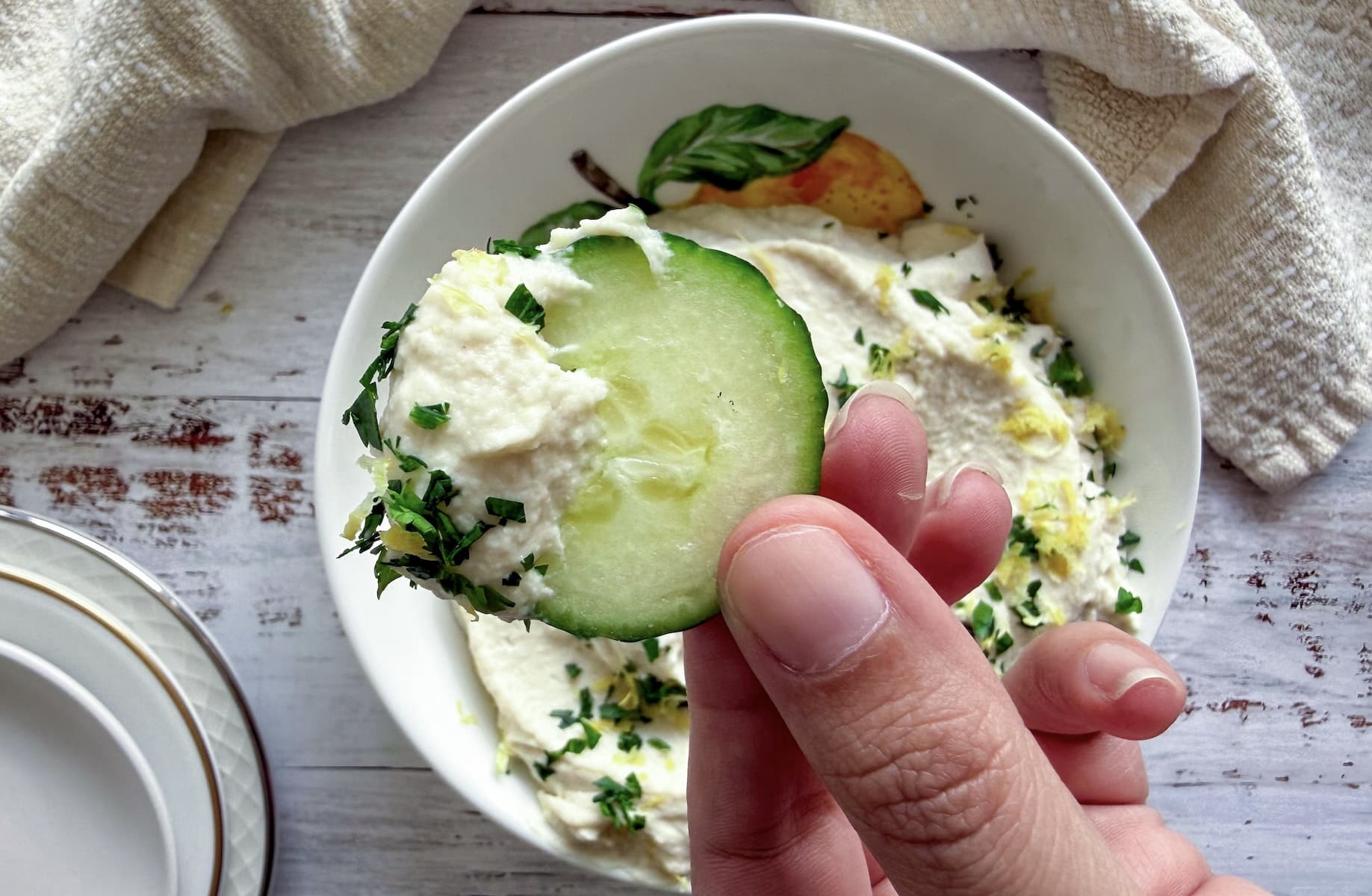 hand with a cucumber with hummus on it over a bowl with more.