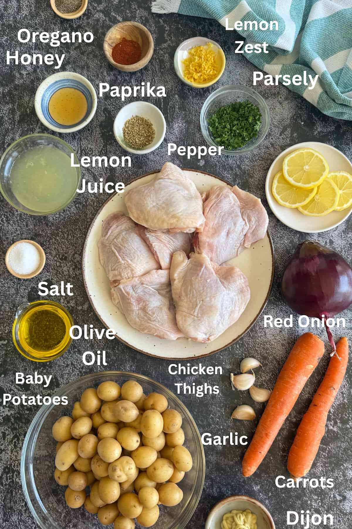 ingredients for chicken and lemon tray bake laid out in small bowls - oregano, honey, paprika, lemon juice, lemon zest, parsley, salt, pepper, red onion, garlic, carrots, olive oil, chicken thighs, baby potatoes, and dijon.