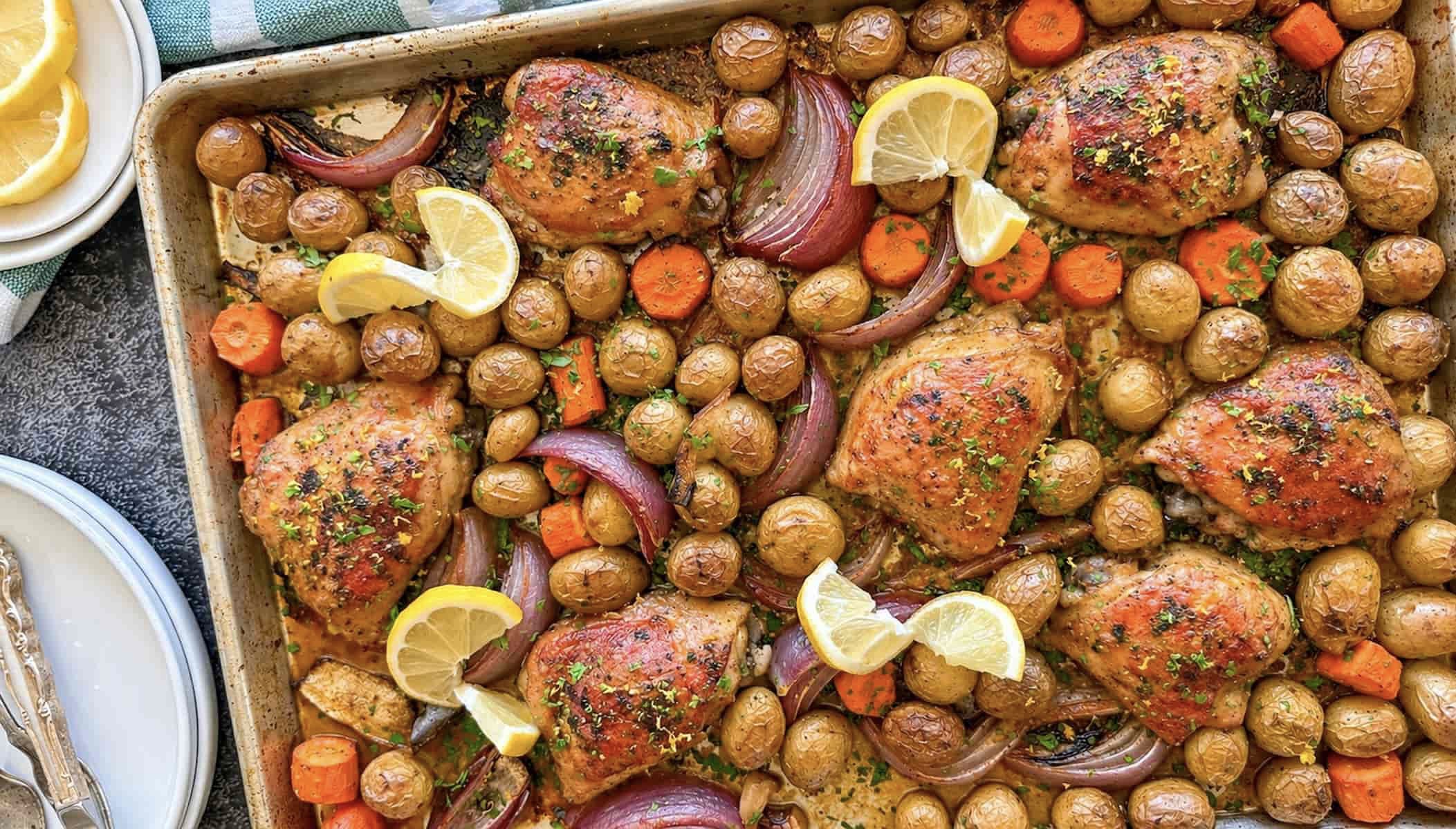 golden chicken thighs on a sheet pan with baby potatoes, carrots, red onion, and lemon slices.