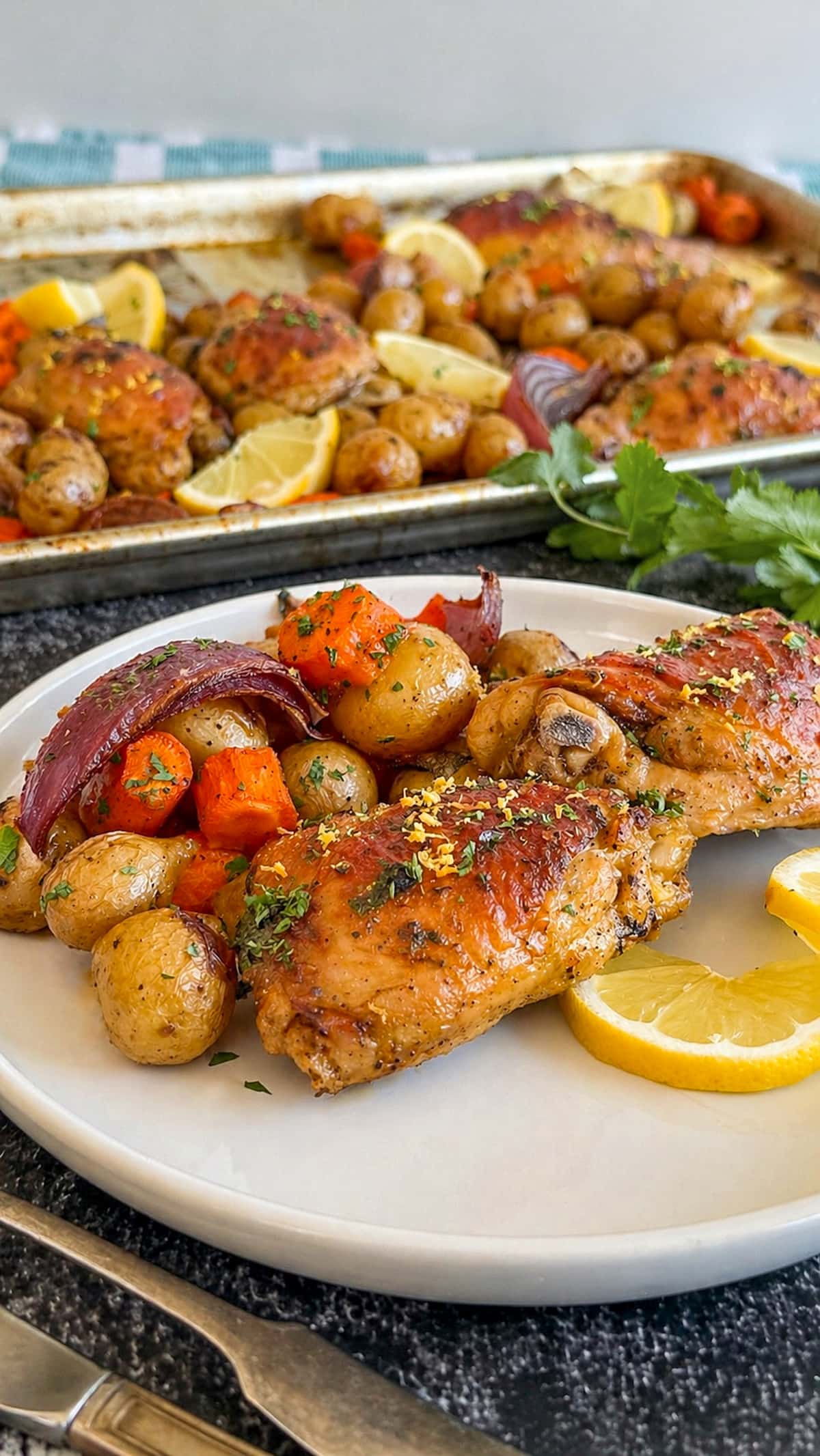 two cooked chicken thighs on a plate with potatoes, red onion, carrots, and lemon. A tray with more in the background.