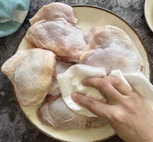 hand with a paper towel patting raw chicken thighs down on a plate.