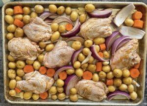 raw seasoned chicken thighs nestled into baby potatoes, carrots, and red onion on a sheet pan ready for the oven.