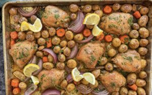 cooked golden chicken thighs on a sheet pan with baby potatoes, carrots, red onion, parsley, and lemon slices.