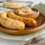 2 cooked chicken sausage on a plate with a small bowl of sauerkraut. 2 more sausages on an air fryer basket in the background.