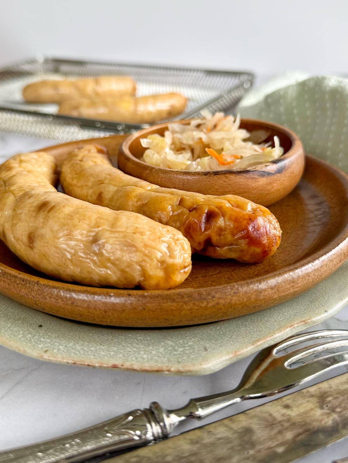2 cooked chicken sausage on a plate with a small bowl of sauerkraut. 2 more sausages on an air fryer basket in the background.