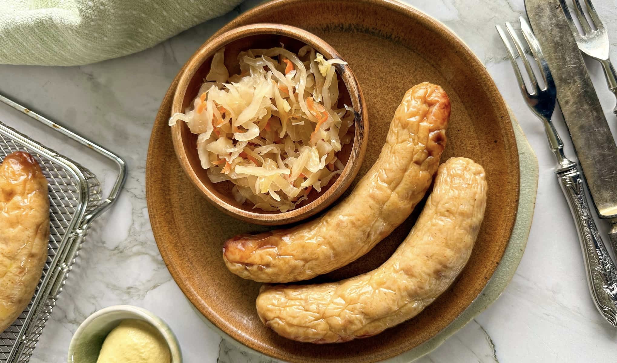2 chicken sausages on a brown plate next to a small bowl of sauerkraut. forks, knives, dijon mustard and the air fryer basket with more sausage in the background.