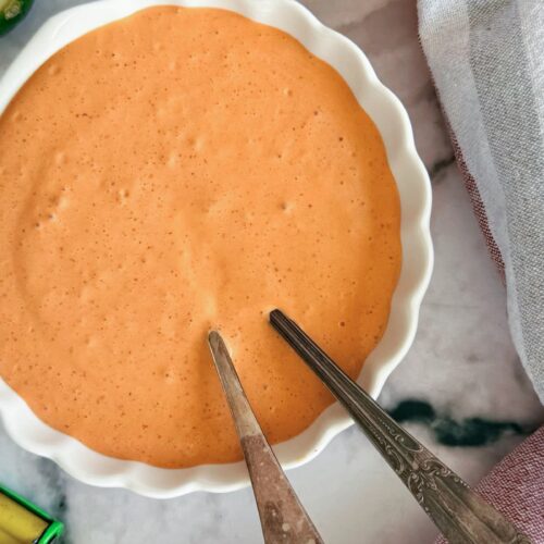 orange chipotle sour cream sauce in a bowl with two spoons. Halved lime, citrus juicer, and can of chipotle peppers in adobo surrounding.