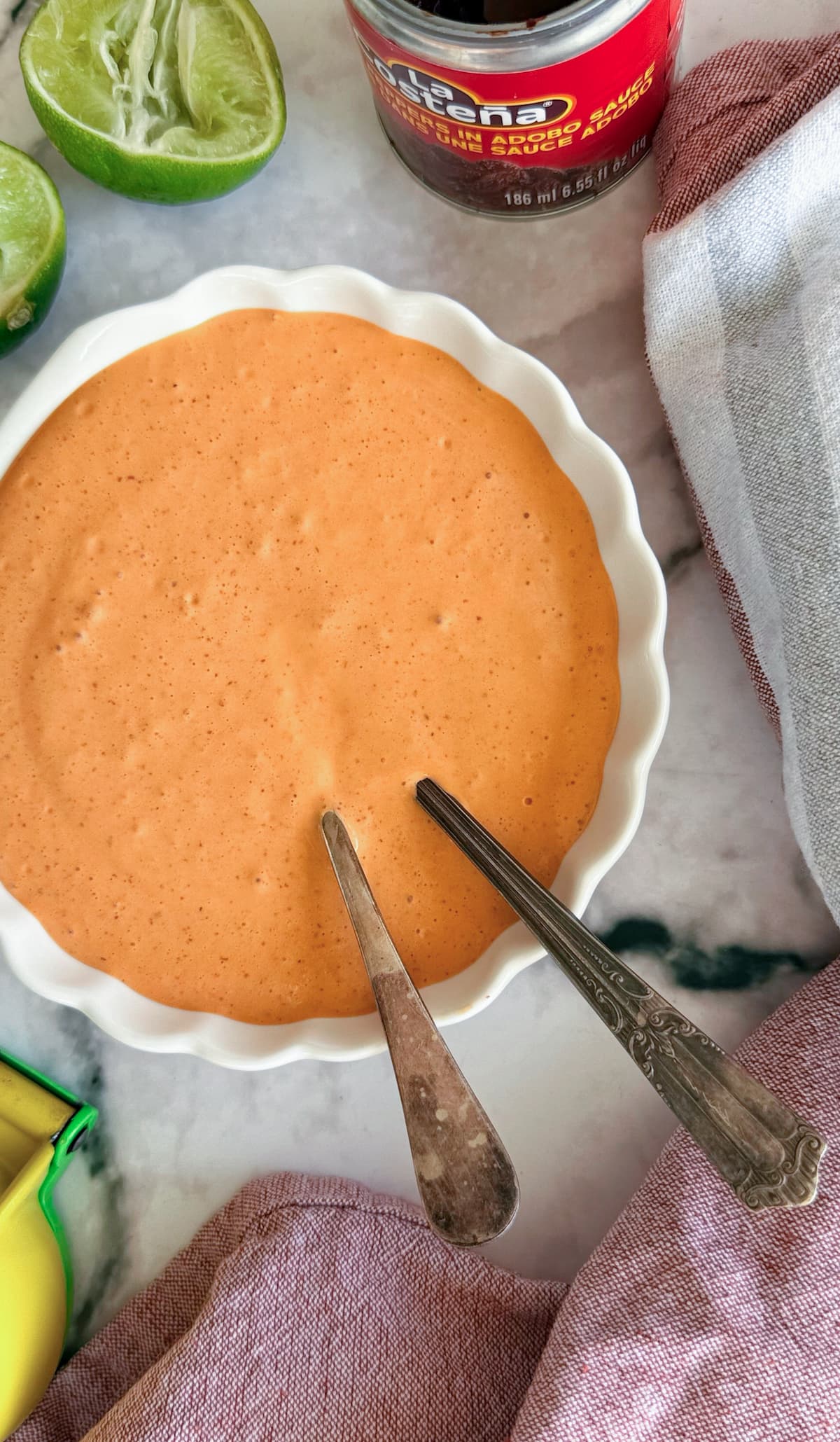 orange chipotle sour cream sauce in a bowl with two spoons. Halved lime, citrus juicer, and can of chipotle peppers in adobo surrounding.