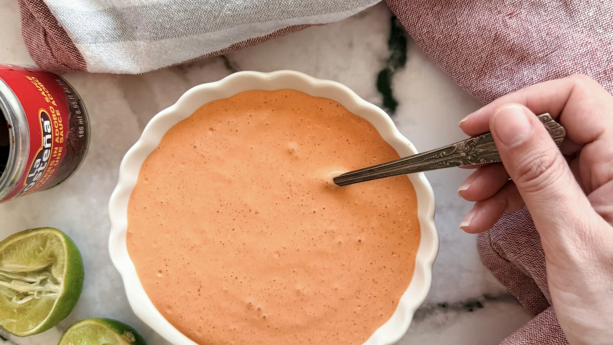 hand holding a spoon in a bowl of an orange chipotle sauce in a white bowl. Halved lime and can of chipotle peppers in the background.